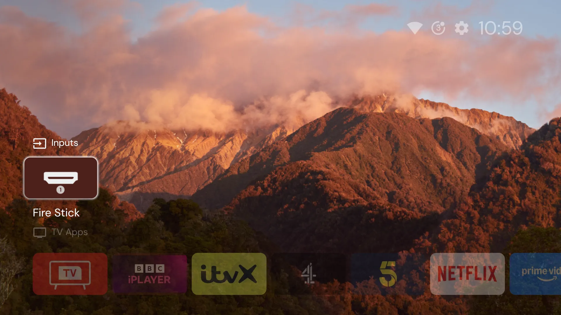 Projectivy’s home screen on the TV: an Inputs row with one tile, Fire Stick, in focus, and below it a row of apps starting with TV, BBC iPlayer, ITVX, Channel 4, 5, Netflix and Prime Video, over a photograph of a mountain at sunset.